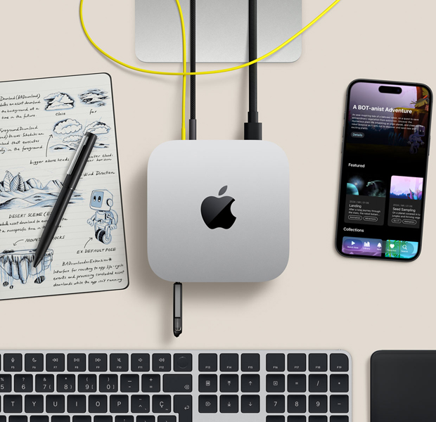 Mac mini, vue en plongée, aluminium argent, coins arrondis, logo Apple noir brillant centré, branché à un câble, clé USB insérée dans le port USB-C à l’avant, support du Studio Display partiellement visible, iPhone, Magic Trackpad, Magic Keyboard avec Touch ID, stylet et carnet d’esquisses à encre électronique