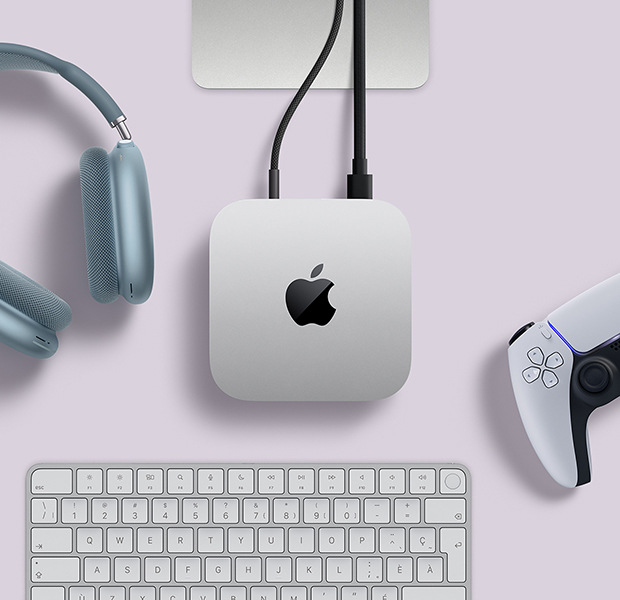 Mac mini, vue en plongée, aluminium argent, coins arrondis, logo Apple noir brillant centré, un câble branché, support du Studio Display partiellement visible, AirPods Max 2, bleu, manette DualSense pour PlayStation, blanche, et clavier blanc