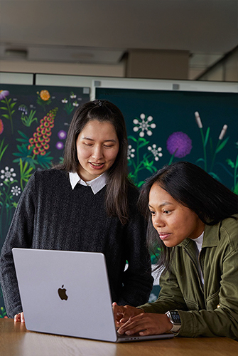 Twee teamleden van Apple Marketing die samenwerken op een MacBook. Ze zitten voor posters die hun team heeft ontworpen.