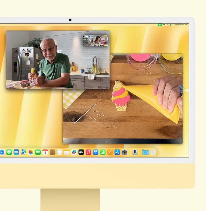 Žlutý iMac. Ukázka funkce Pohled na stůl. Na obrazovce je cukrářka při videohovoru s další osobou. V jednom okně se zobrazuje cukrářka a tvář muže. Ve druhém je vidět ruka cukrářky při zdobení sladkého pečiva.