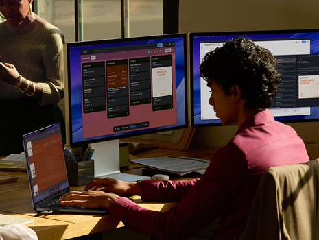 A team of employees sitting around a table in an office setting. A woman sits at her desk working on a MacBook and displays. Her colleagues are gathered around in discussion.