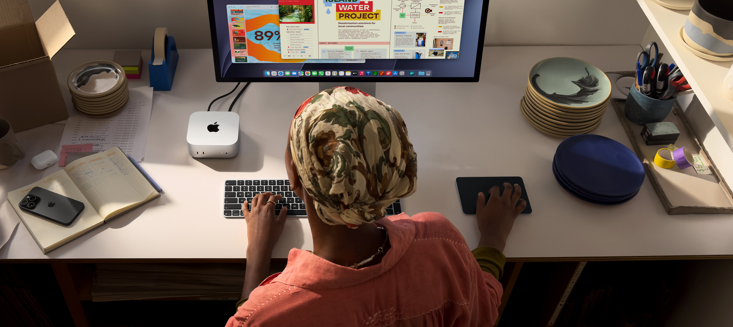 Íróasztalnál ülő ember egy Mac minin gépel, melyhez Studio Display, Magic Keyboard és Magic Trackpad van csatlakoztatva, körülötte különféle tárgyak