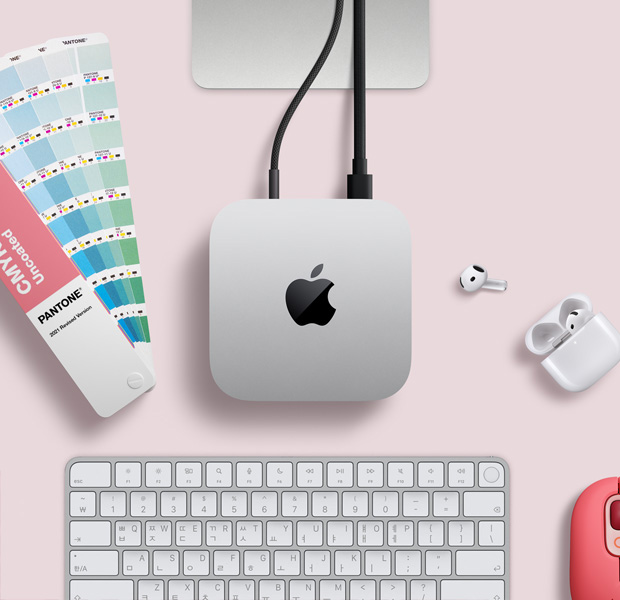 Mac mini를 위에서 바라본 모습. 실버 알루미늄 외장, 둥근 형태의 모서리, 중앙의 광택 나는 검은색 Apple 로고가 보이며, 코드가 연결되어 있습니다. Studio Display의 스탠드 일부가 보이고, AirPods 4, 팬톤 색상 견본, 빨간색 Logitech 키보드와 마우스가 놓여 있습니다.