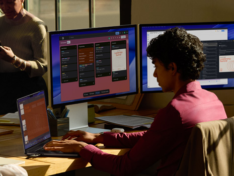 A team of employees sitting around a table in an office setting. A woman sits at her desk working on a MacBook and displays. Her colleagues are gathered around in discussion.