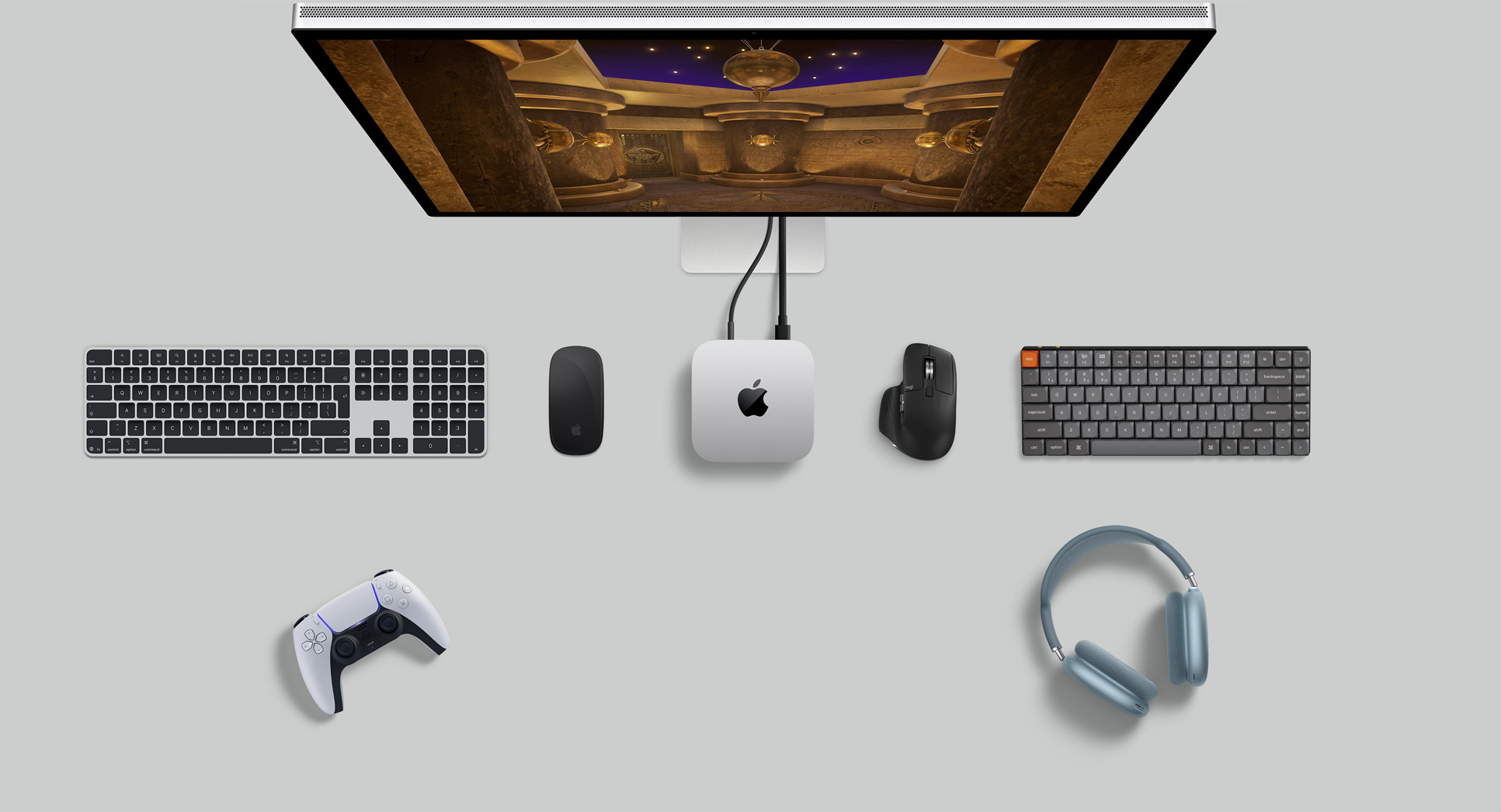 Top view of Mac mini in a desk setup with Studio Display, wireless keyboards, wireless mice, a gaming controller, and AirPods Max