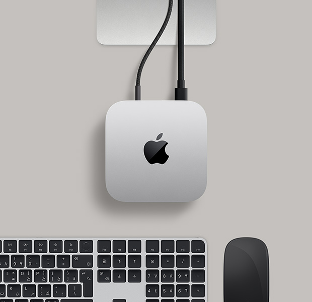 جهاز Mac mini، الجهة العلوية، ألومنيوم فضي، زوايا مدورة، شعار Apple باللون الأسود اللامع في الوسط، قاعدة شاشة Studio Display تظهر بشكل جزئي، لوحة مفاتيح ماجيك مع ميزة بصمة الإصبع، لوحة تعقب ماجيك‏