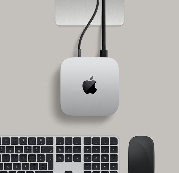 Vista desde arriba, Mac mini, aluminio color plata, esquinas redondeadas, logo negro brillante de Apple en el centro, base de un monitor Studio Display parcialmente visible, Magic Keyboard con Touch ID, Magic Trackpad