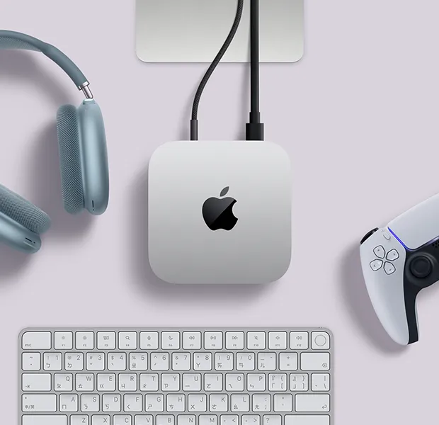 Mac mini 的俯視圖，展示銀色鋁金屬機身、圓角設計、機身中央的黑色亮面 Apple 標誌，透過連接線連接，Studio Display 支架部分可見，旁邊放著藍色 AirPods Max 2、白色 PlayStation DualSense 控制器和白色鍵盤