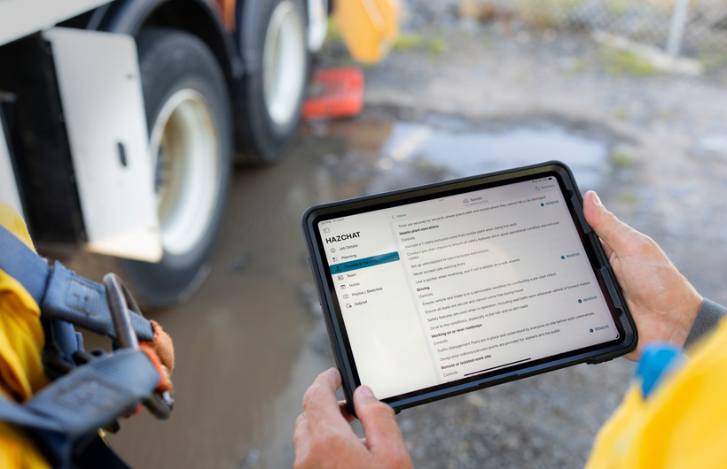 Unas manos sostienen un iPad con la app HazChat abierta que muestra Datos del trabajo, Planificación, Peligros y Controles, en un lugar de trabajo junto a un charco de barro