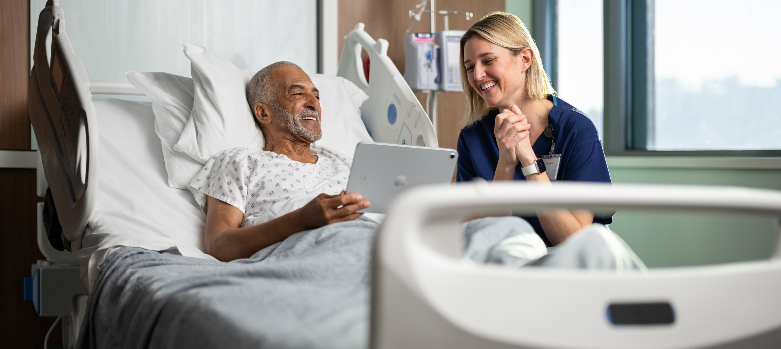 En mandlig patient sidder i en hospitalsseng. Han smiler, mens han kigger på en iPad-skærm sammen med en kvindelig sygeplejerske.