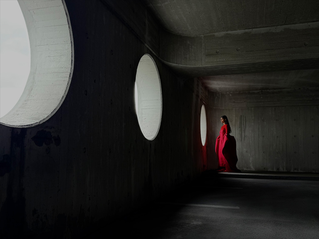 Eine Frau in einem langen roten Kleid steht in einem leeren Raum und wird fotografiert. Die Fotos App wechselt durch die Brennweiten, um die Möglichkeiten des Pro Kamera-Systems zu zeigen, hier: 35 mm