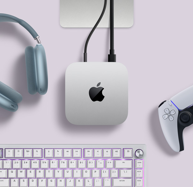 Mac mini, top, silver aluminum, rounded corners, glossy black Apple logo centered, connected by cord, Studio Display stand partially visible, AirPods Max 2, blue color, PlayStation DualSense controller, white color, and white keyboard